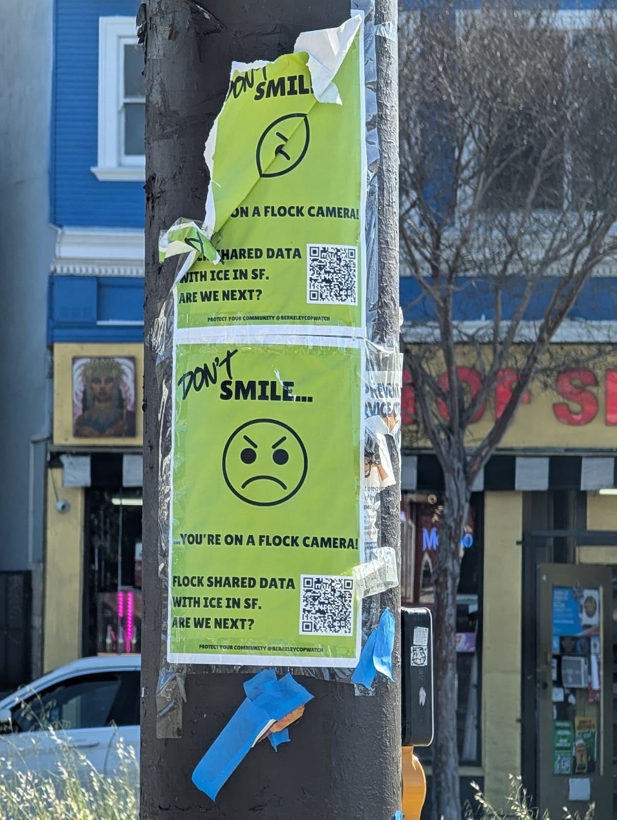 A day-glo yellow sign with an angry-faced emoticon. It reads, "DON'T SMILE...YOU'RE ON A FLOCK CAMERA! FLOCK SHARED DATA WITH ICE IN SF. ARE WE NEXT?"