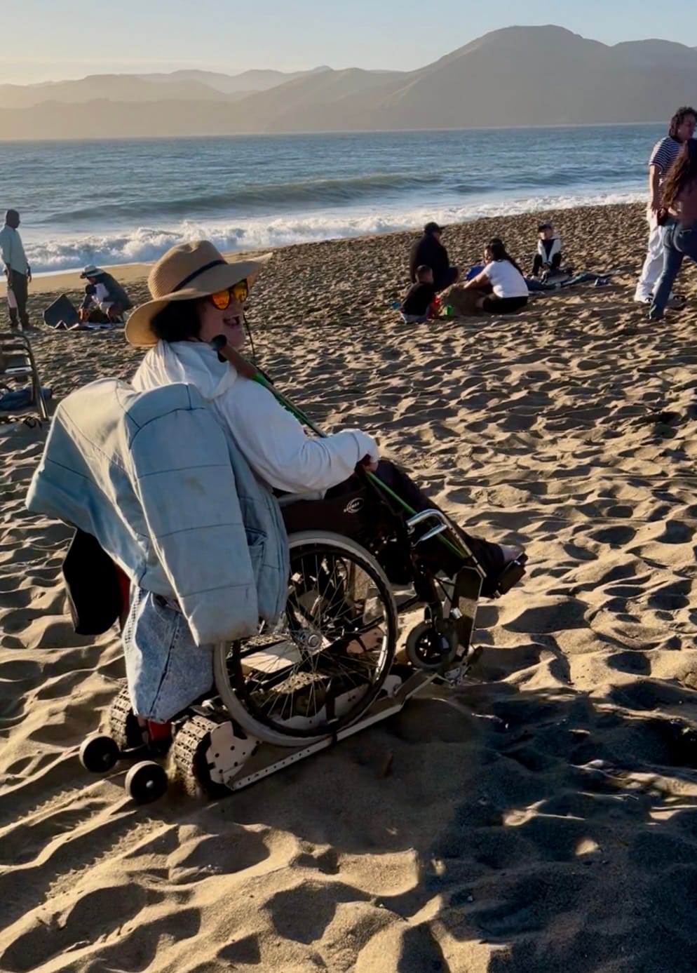 A smiling woman in her power chair on some sand by the ocean, late afternoon sun behind her, people hanging around nearby.