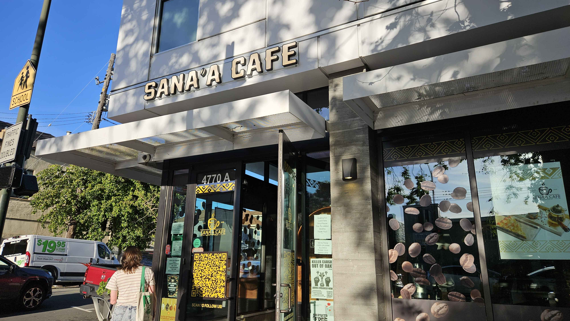 The exterior of a new construction building, the storefront of Sana'a Cafe. The door is open, and the windows are filled with signs, political posters, static cling-ons of coffee beans, geometric designs, QR codes, etc.