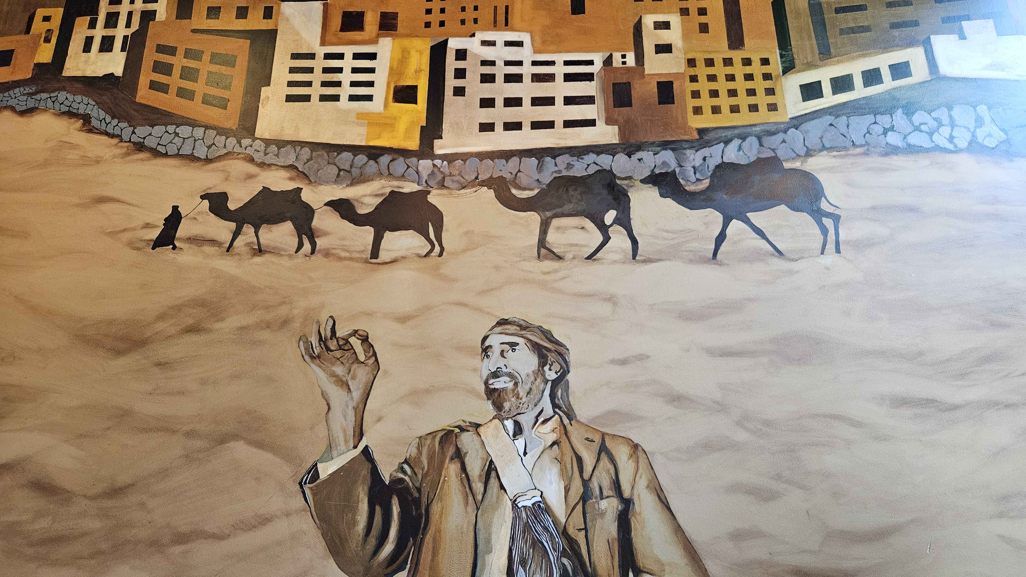A wall painting. In the foreground, a bearded man with a traveler's bag inspects a single coffee bean with a pleased expression. Behind him, the sillhouettes of a person leading a team of four camels. Behind them, a stone wall surrounding a dense city skyline.