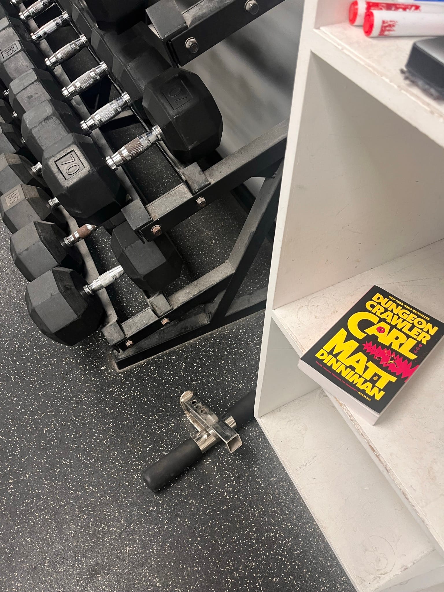 A copy of "Dungeon Crawler Carl" on a shelf in a gym next to a rack of dumbbells.