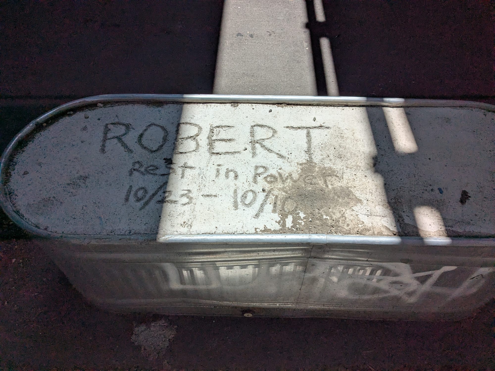 "Robert Rest in Power 10/23-10/10" written in the concrete of a planter used as anti-unhoused hostile architecture