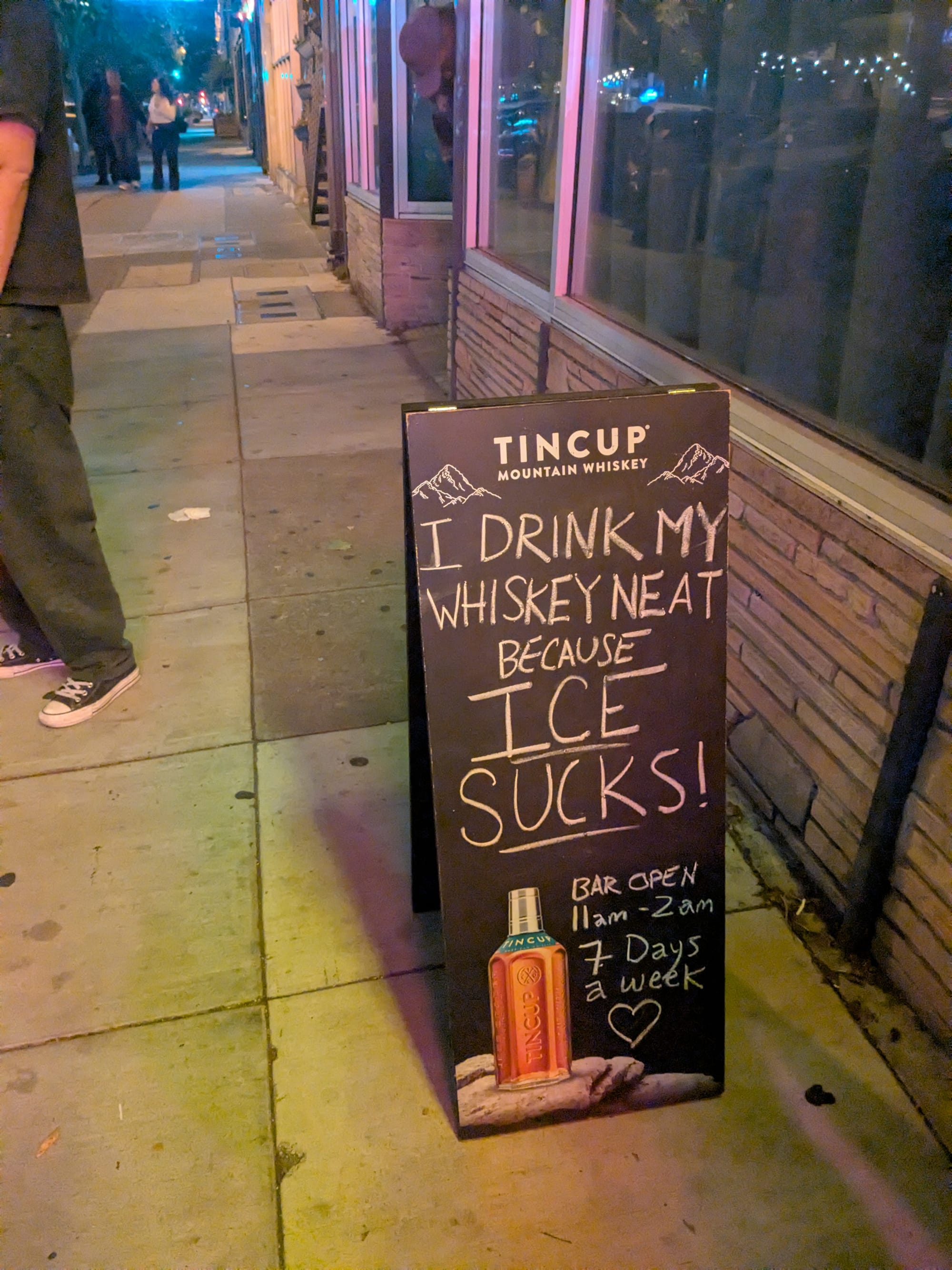 A sign for a bar on the street that reads "I drink my whiskey neat because ICE SUCKS"