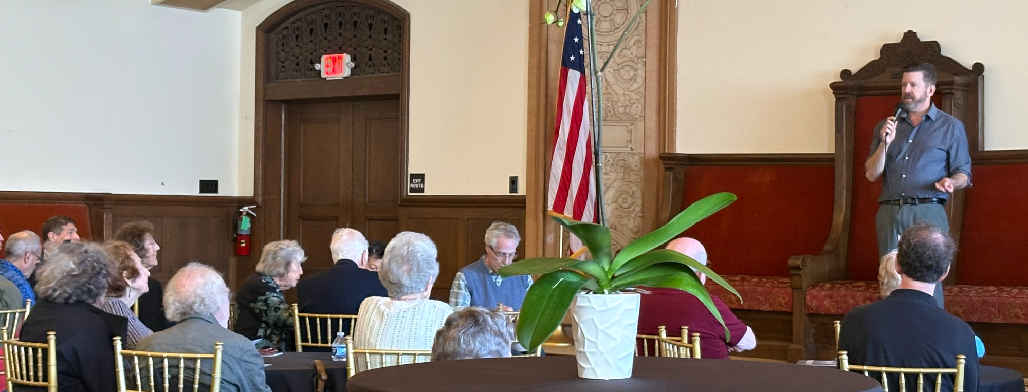 A man standing in front of an ornate chair talking to a room full of people demographically clustering towards their golden years.