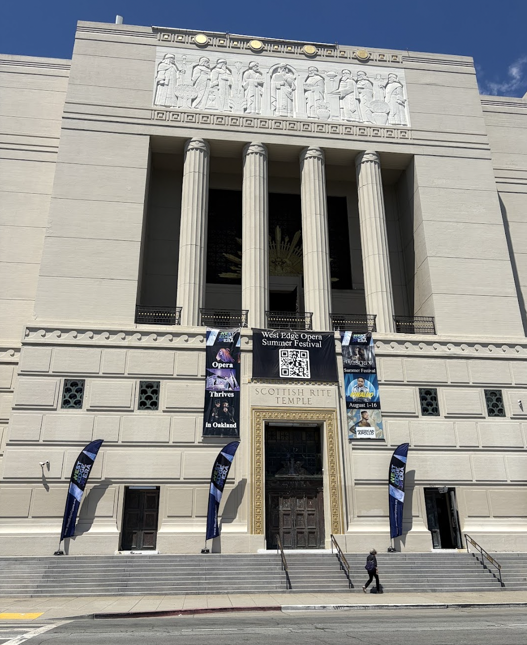 Exterior of the kind of fancy ass building that would be called a "Scottish Rite Temple" with banners for West Edge Opera, in bright sunlight.