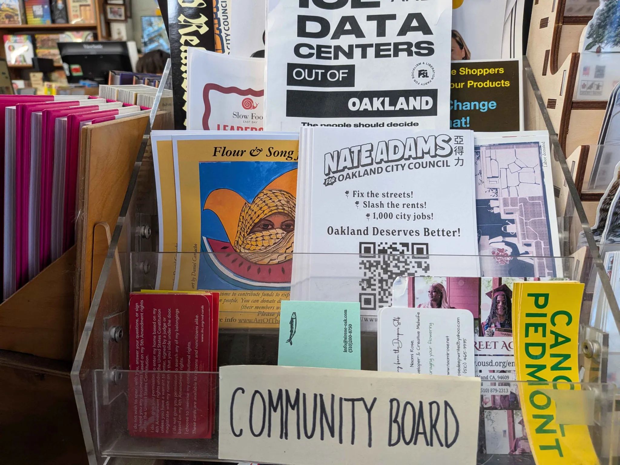 A photo of a Nate Adams flyer at a bookstore, situated in the "community board" among "CANCEL PIEDMONT" stickers and a "DATA CENTERS OUT OF OAKLAND" flyer.