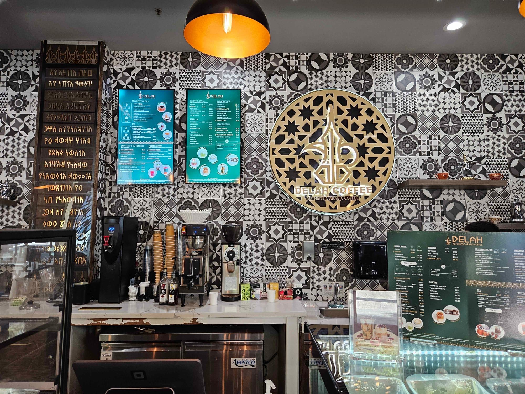 A view of the counter at Delah Coffee. The wall is decorated with an array of black & white geometric tiles. Ancient Southern Arabian script lines a column next to various coffee machines.