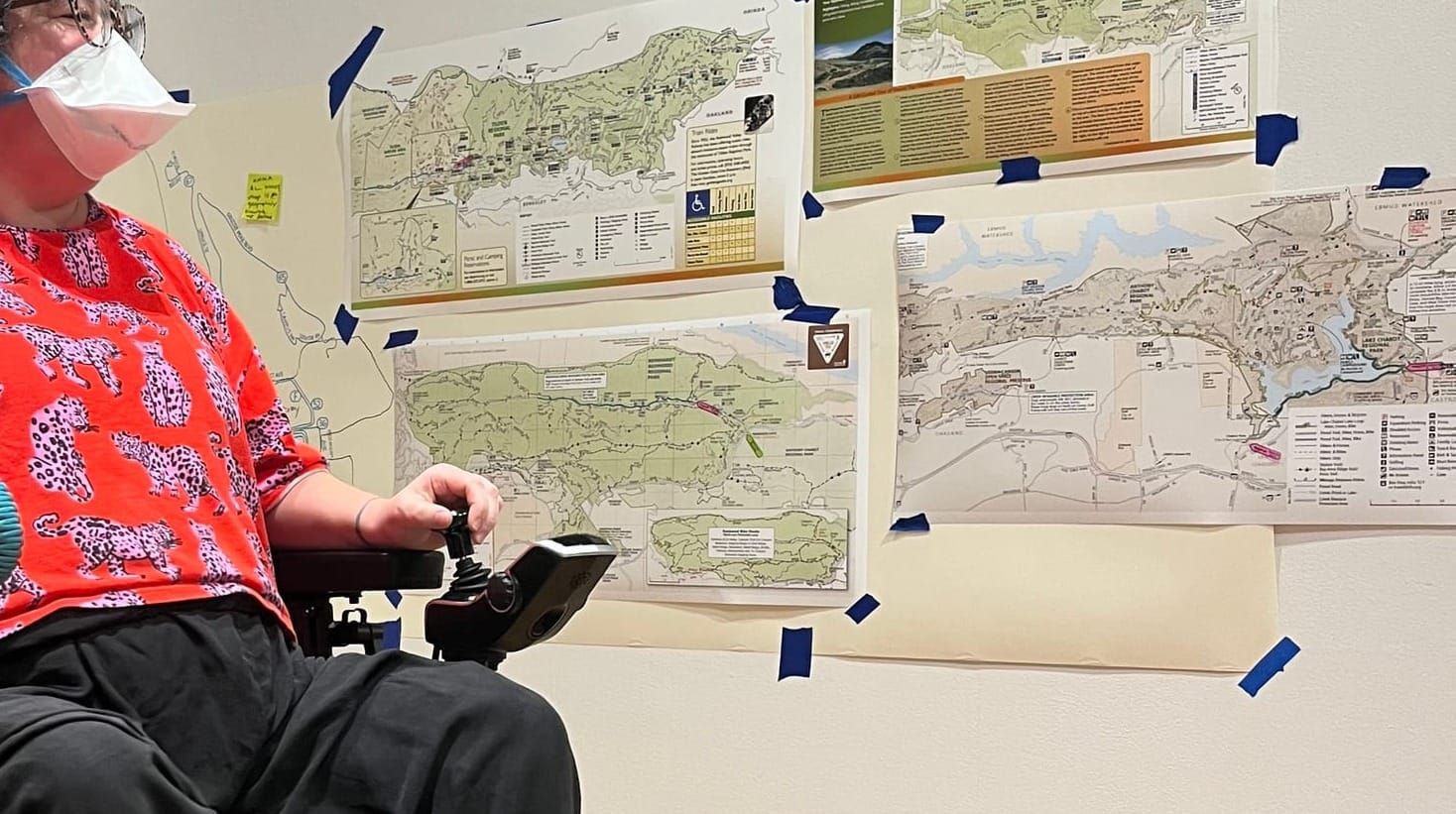 A woman wearing a colorful top printed with tigers moves her power chair next to a wall hung with maps of East Bay Parks.
