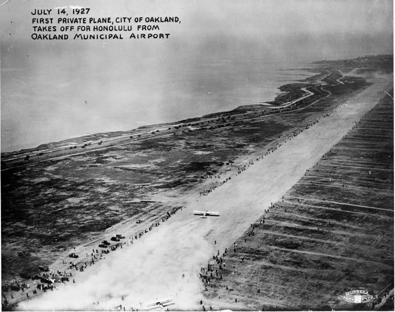 A black-and-white photo showing a strip of runway with people lining it on either side. The bay is on the left.