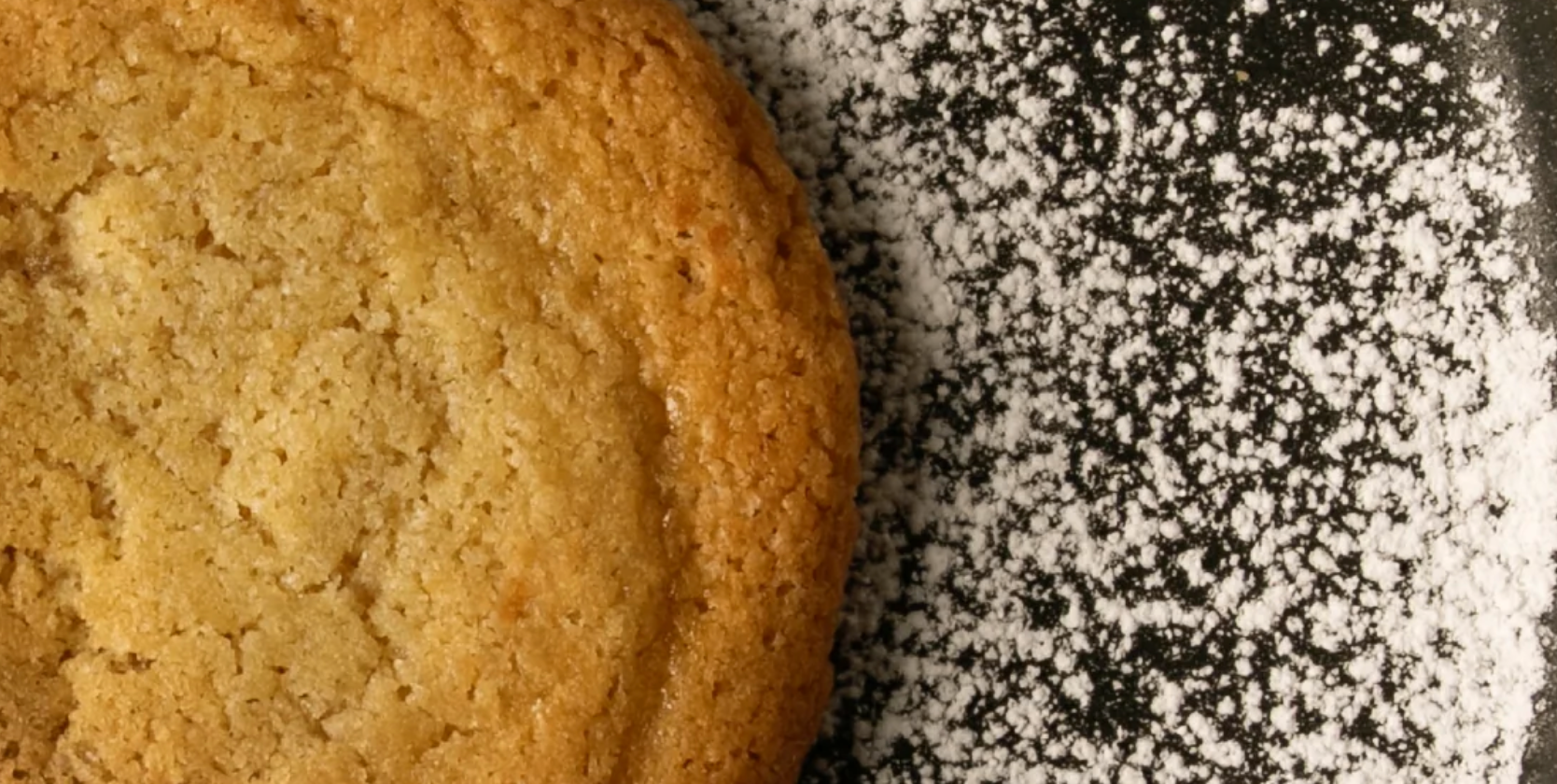 A brown butter cookie and powdered sugar on a dark plate.