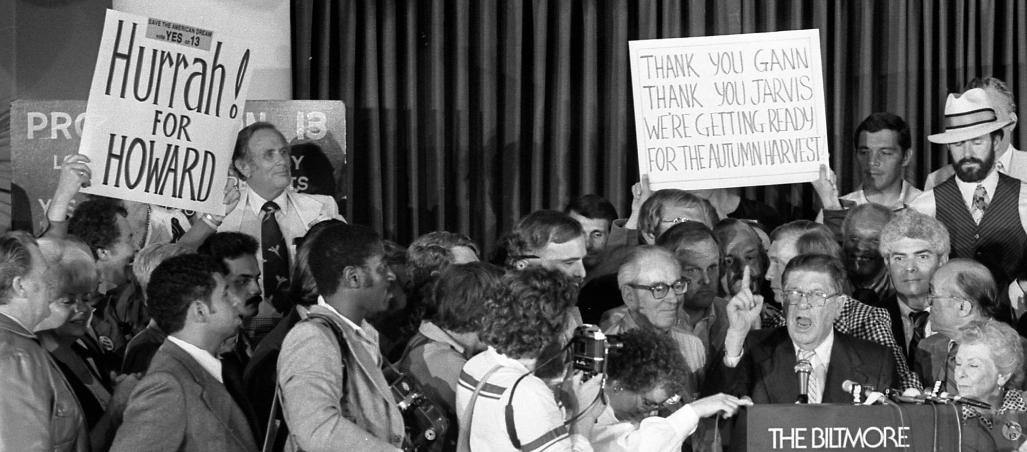Howard Jarvis, that sack of shit, points a finger skyward while standing at a lectern, surrounded by happy incontinent old people. There are dumb signs visible.