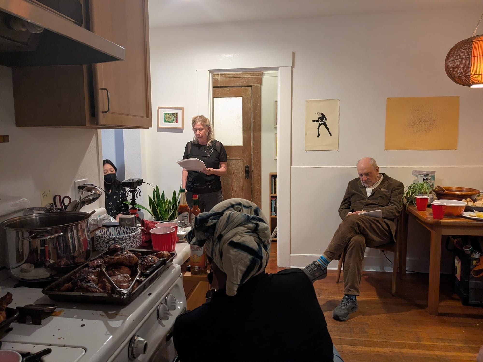 A photo of a warmly appointed Berkeley kitchen, with Spahr reading, Charles Bernstein seated nearby, a couple people listening, some fried chicken on the stovetop.