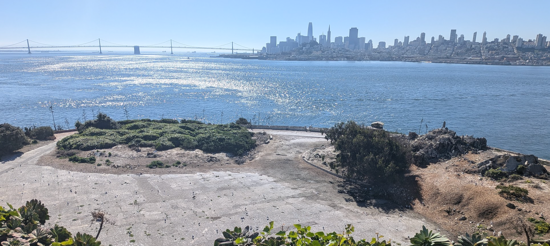 The view of the bay from Alcatraz on a bright sunny day, with the San Francisco skyline and the Bay Bridge visible in the distance.