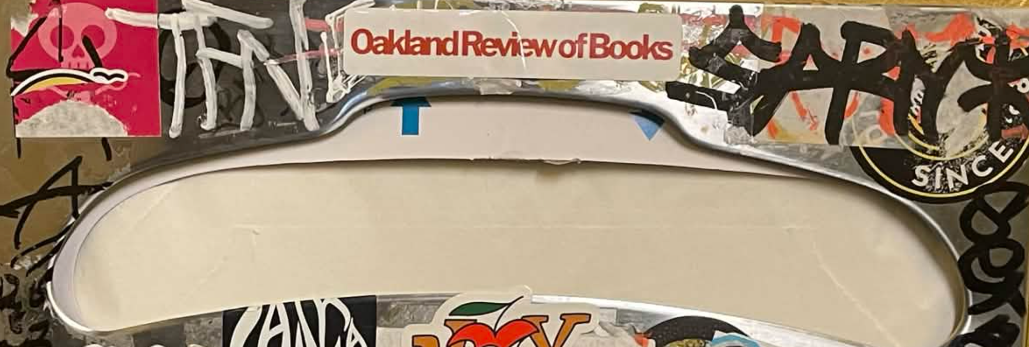 A photo of a toilet seat dispenser in the bathroom of a bar that is NOT Eli's. There's an "Oakland Review of Books" sticker on there, surrounded by graffiti and other stickers. 