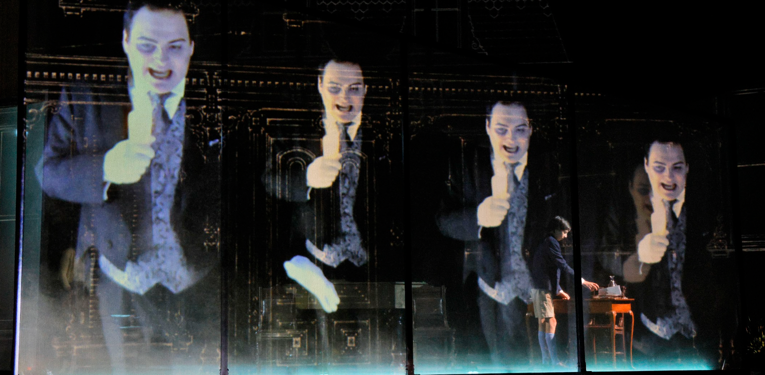 Images of a somewhat unsettling man projected on a screen in front of a boy on stage. 