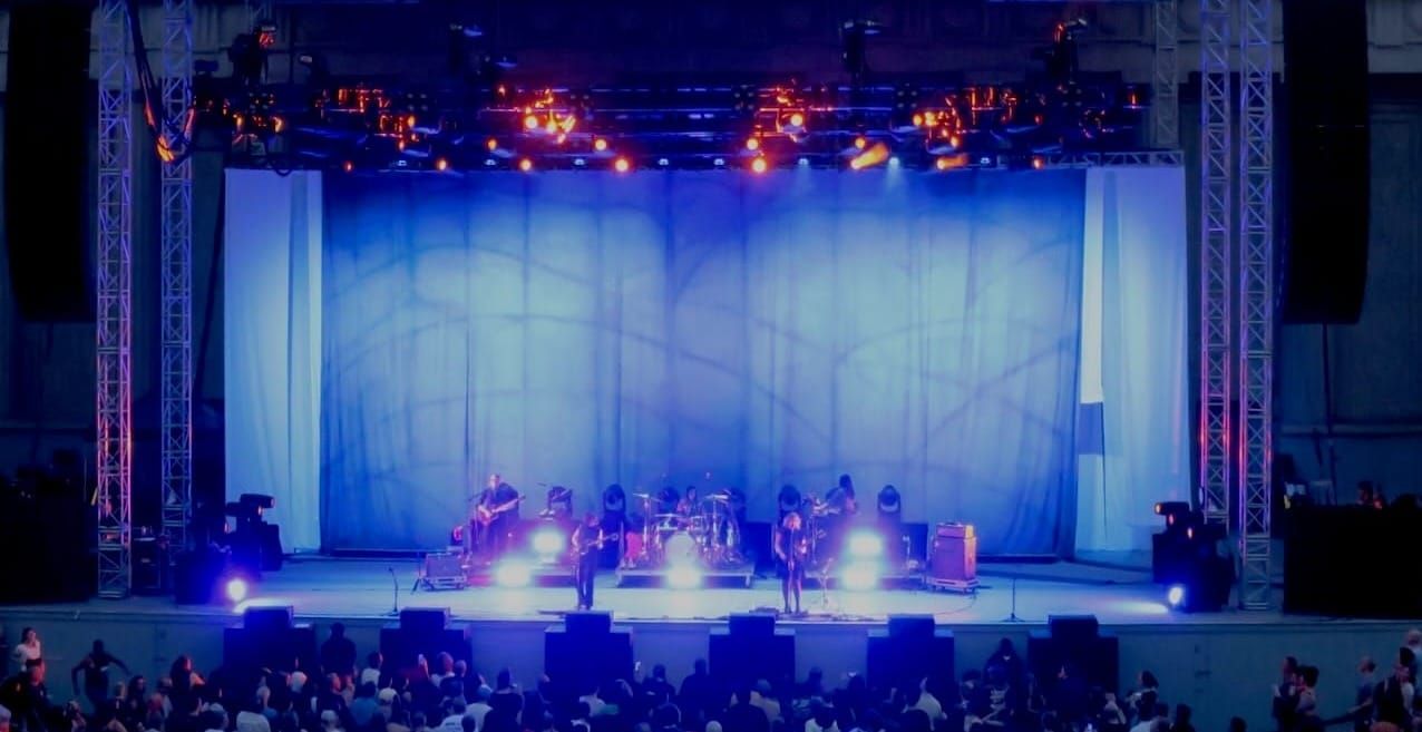 A photo of the stage at the Greek Theater, everything hued blue by the stage lights. Sleater-Kinney is on stage.ey 