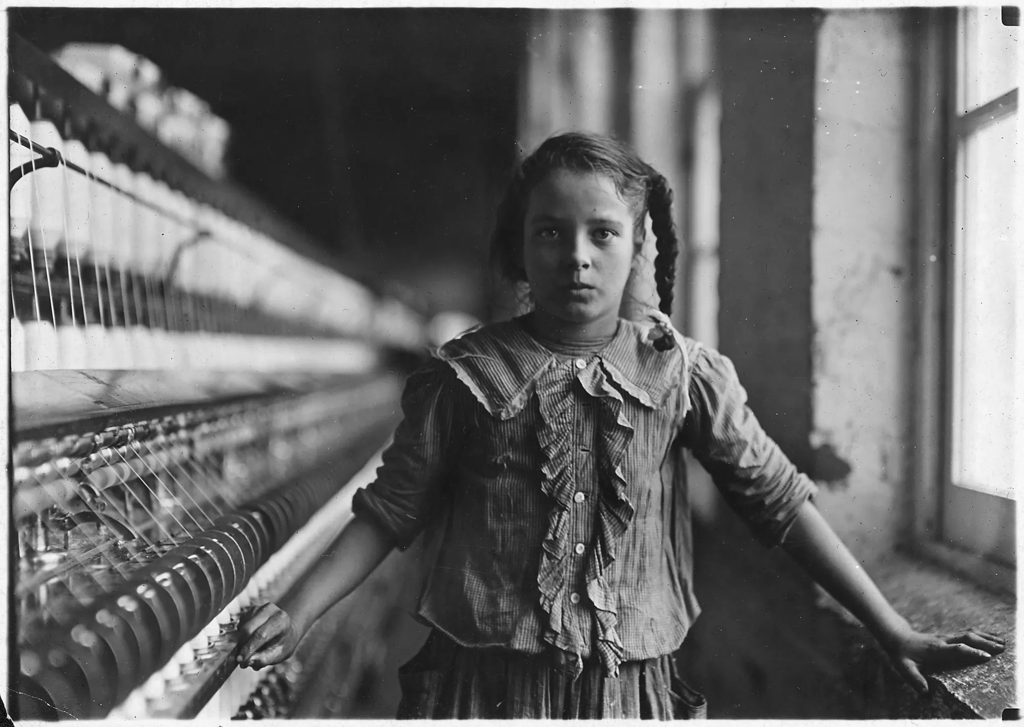 Photograph by Lewis Hine, “One of the spinners in Whitnel Cotton Mill. She was 51 inches high. Has been in the mill one year. Sometimes works at night. Runs 4 sides—48 [cents] a day. When asked how old she was, she hesitated, then said, ‘I don’t remember,’ then confidentially, ‘I’m not old enough to work, but do just the same.’ Out of 50 employees, ten children about her size. Whitnel, N.C.” 12/22/1908. (National Archives Identifier 523145)