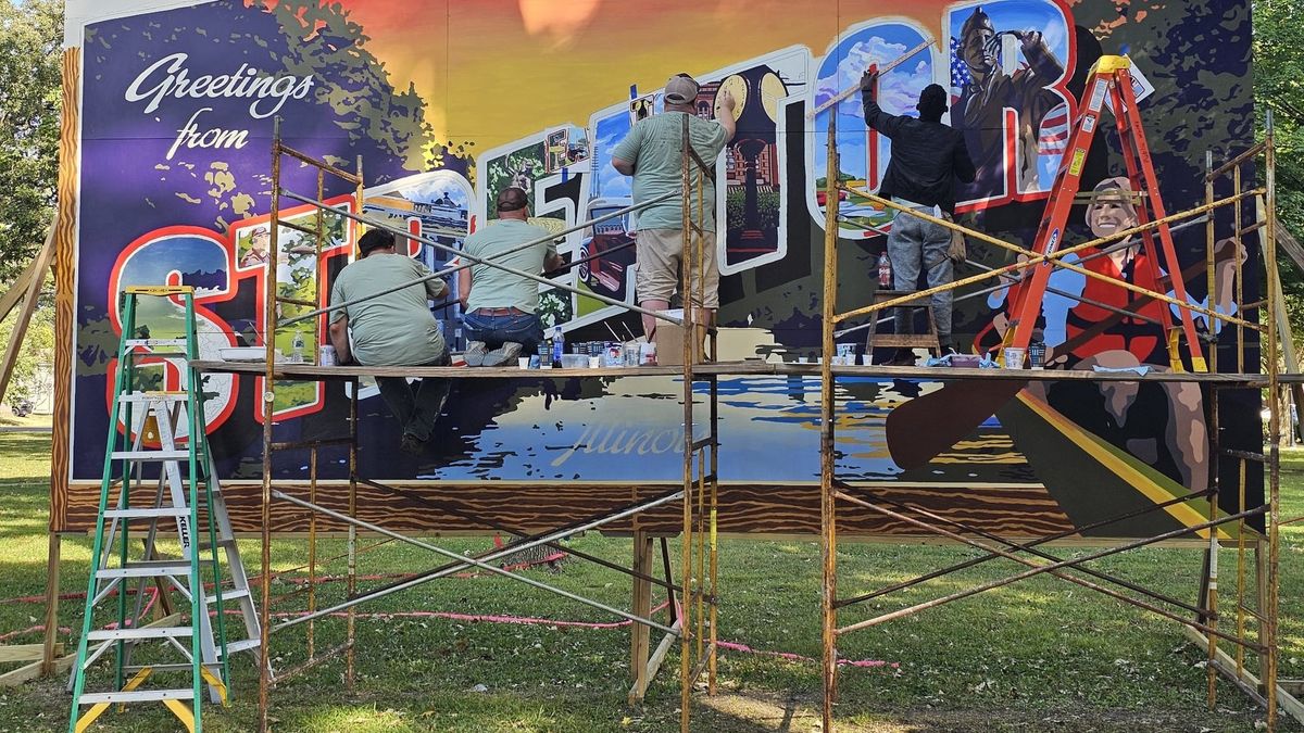 Volunteers on scaffolding painting a Greetings from Streator postcard-style mural in a park