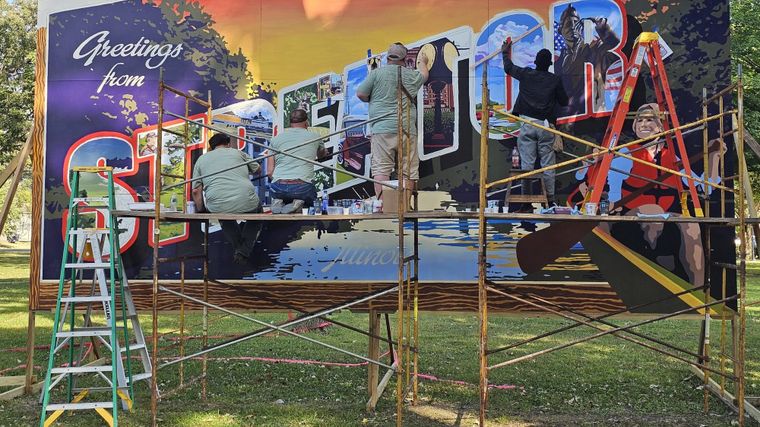 Volunteers on scaffolding painting a Greetings from Streator postcard-style mural in a park