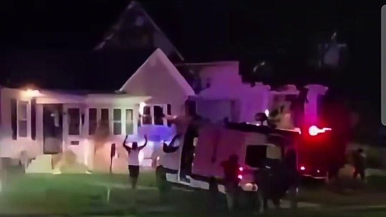 A man with both hands raised walks across a lawn toward an armored truck and agents at night