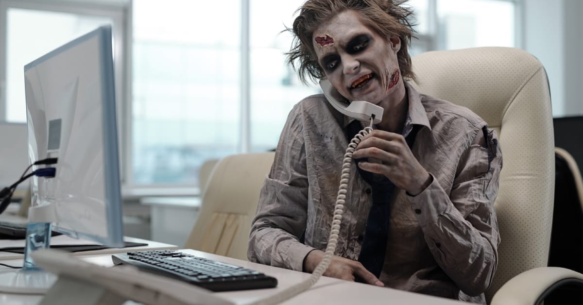Office worker in zombie makeup holding a telephone receiver beside a desktop computer.