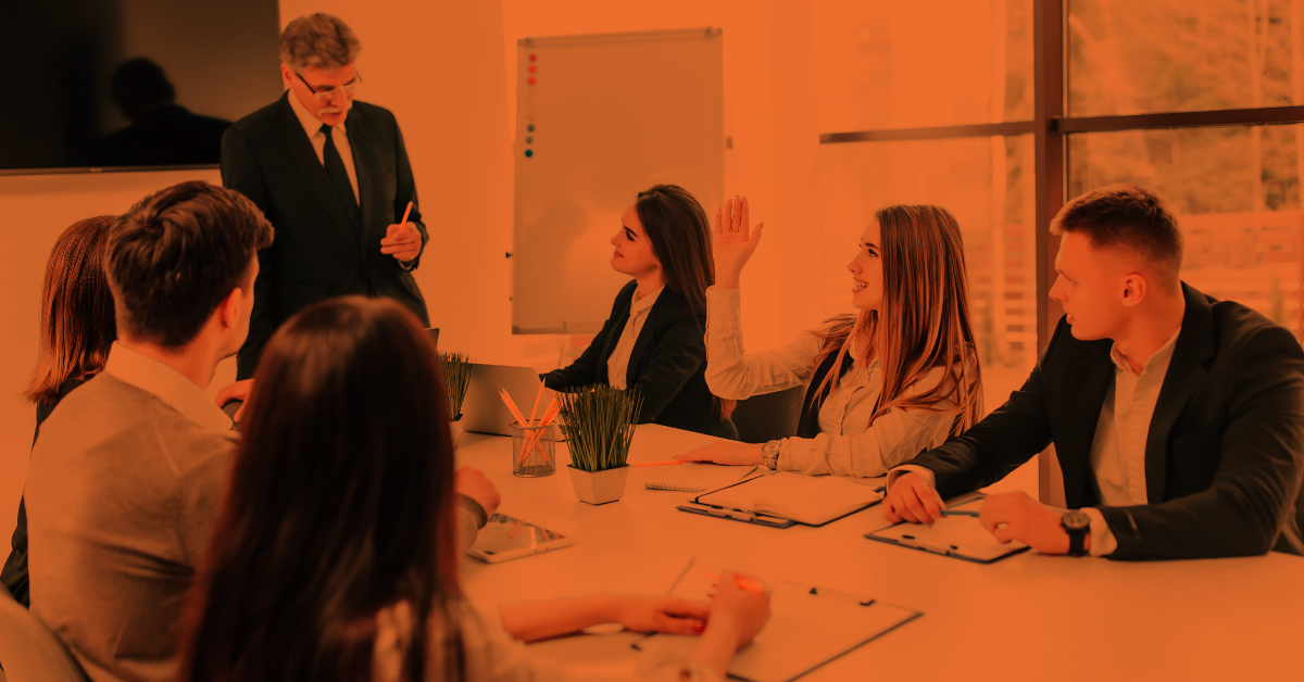 Employees in a meeting and a senior professional speaking while a young woman is raising her hand. 