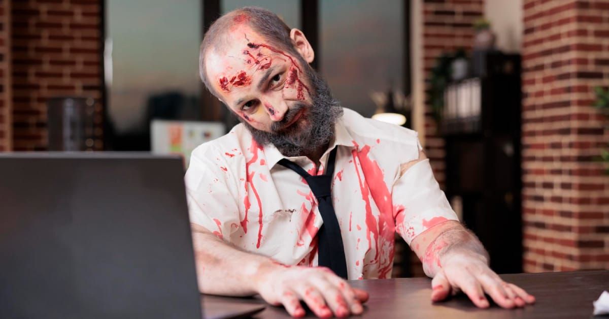Bearded office worker in zombie makeup and a torn, bloodstained costume sitting at a desk with a laptop.
