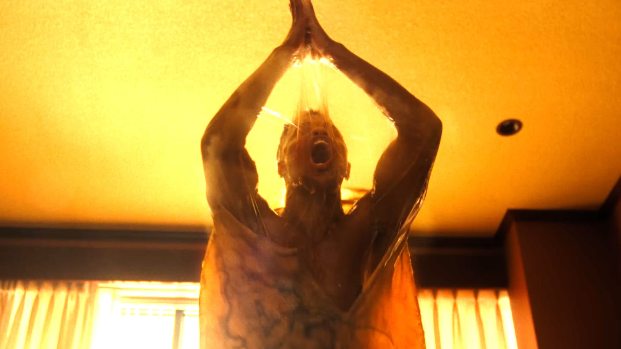 A man emerges from a kind of slime cocoon in an orange-lit room.