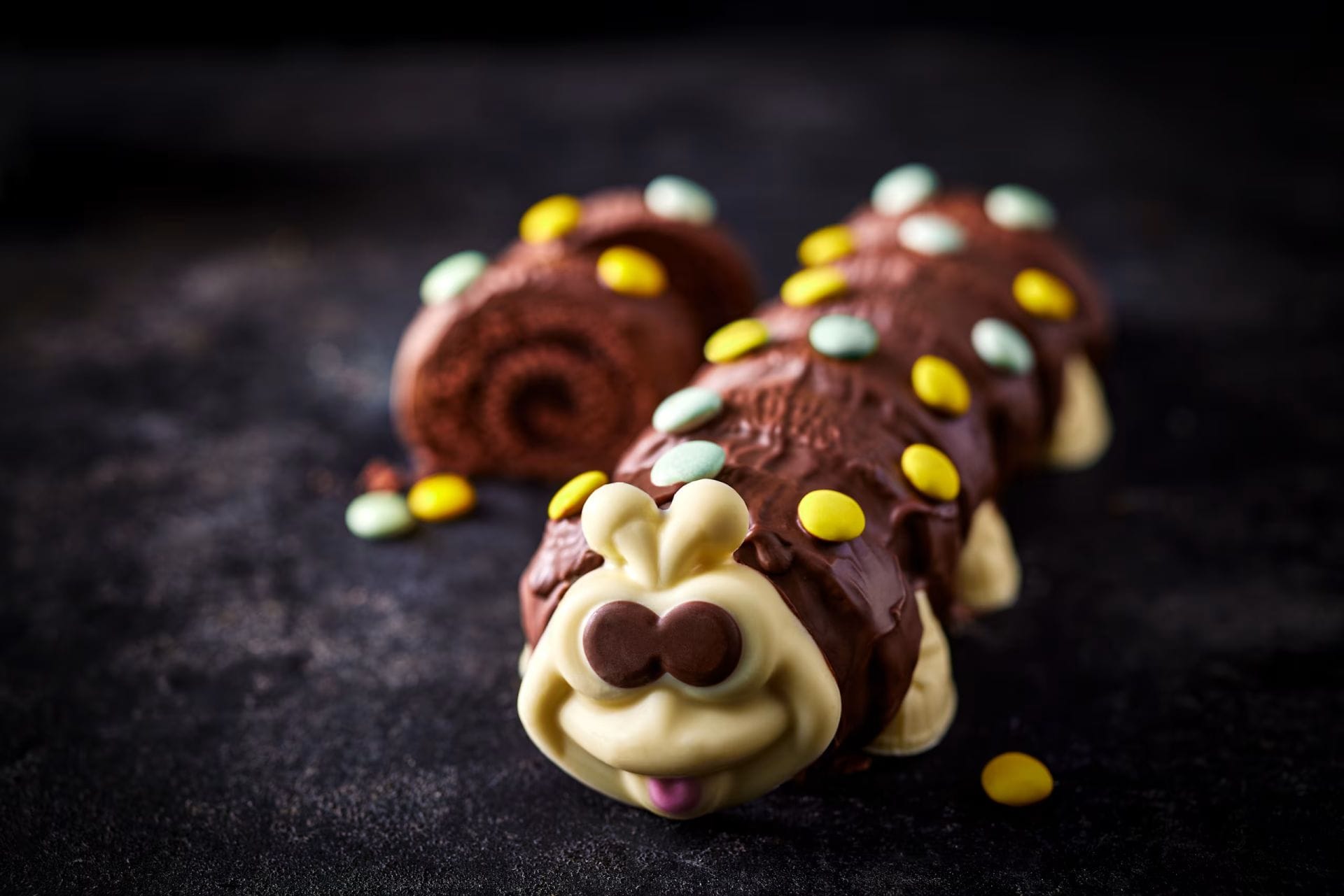 A photo of a Colin the Caterpillar cake from the front, looking at his cute face. A sliced open or rear shot is seen in the background. It's chocolate cake with chocolate topping and a white chocolate face. Smarties are used as decoration.