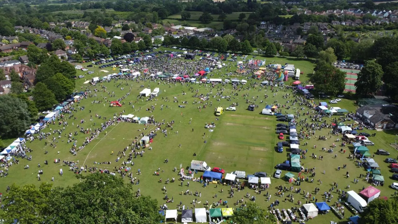 An aerial photo of a summer fair, showing a large field filled with stalls, people, cars, and trees. Houses are seen in the distance surrounding the field.