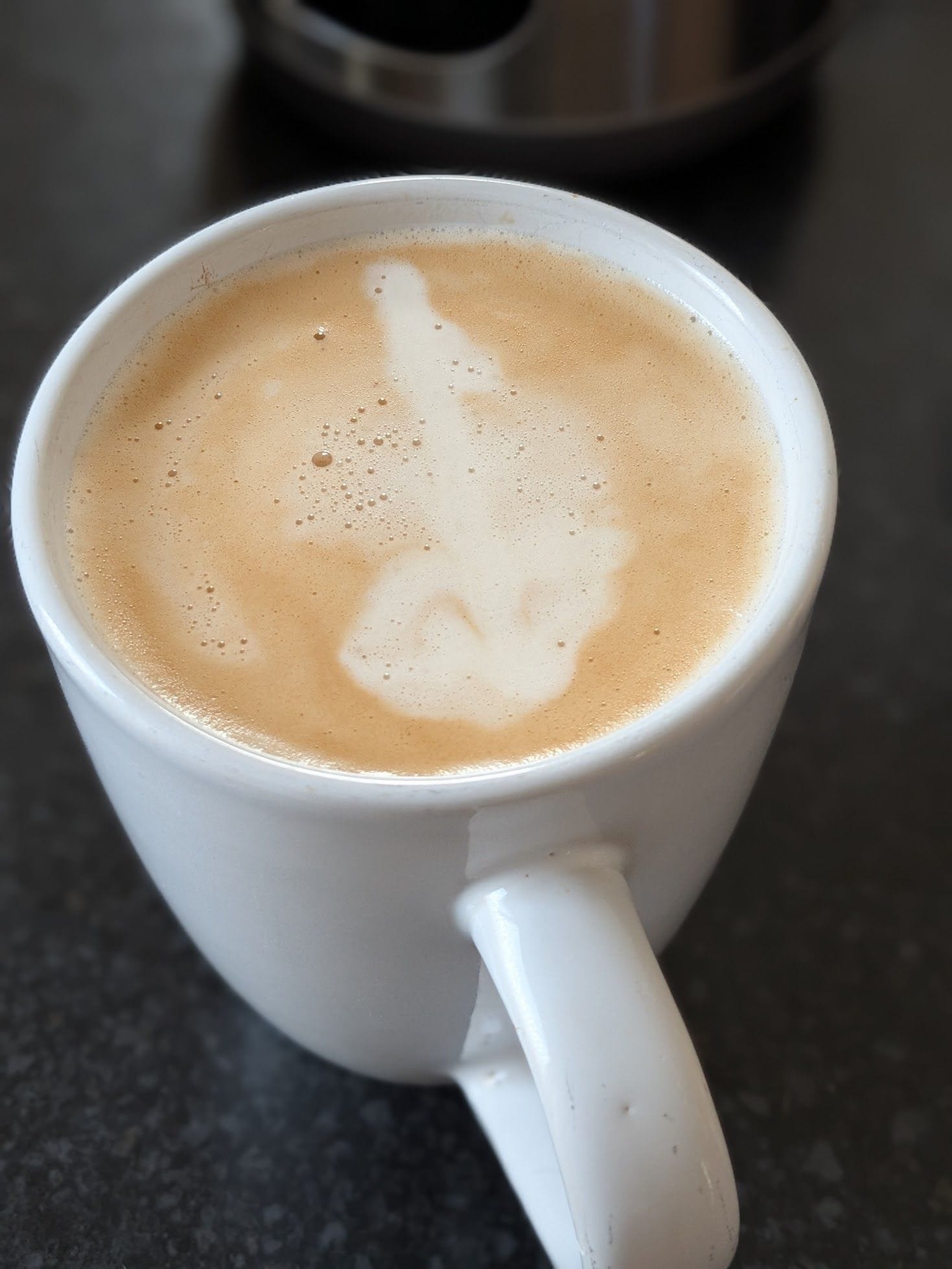 A cup of coffee. The cup is white. The coffee has a pattern on top that looks sort of like a treble clef.