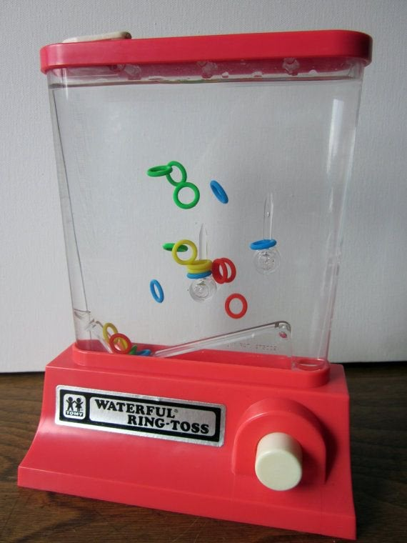 A photo of an old water game, where you press a button on the side to push air into the chamber, moving the rings around and ideally onto the spikes.