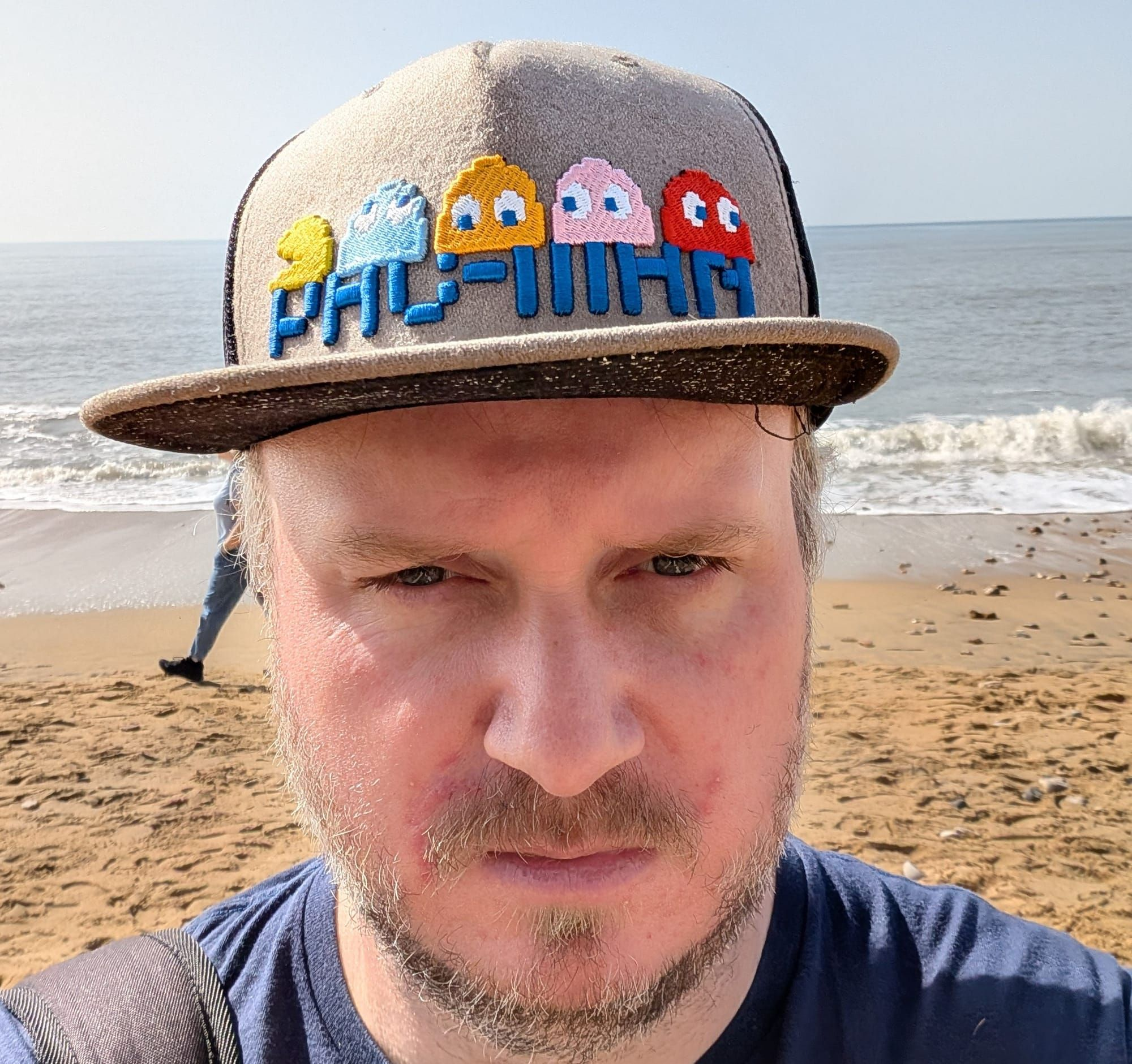 Headshot of Tom Orry, a bearded man wearing a cool Pac-Man baseball cap. He's stood on a sandy beach, the sea visible in the background. A person walks annoyingly behind him.
