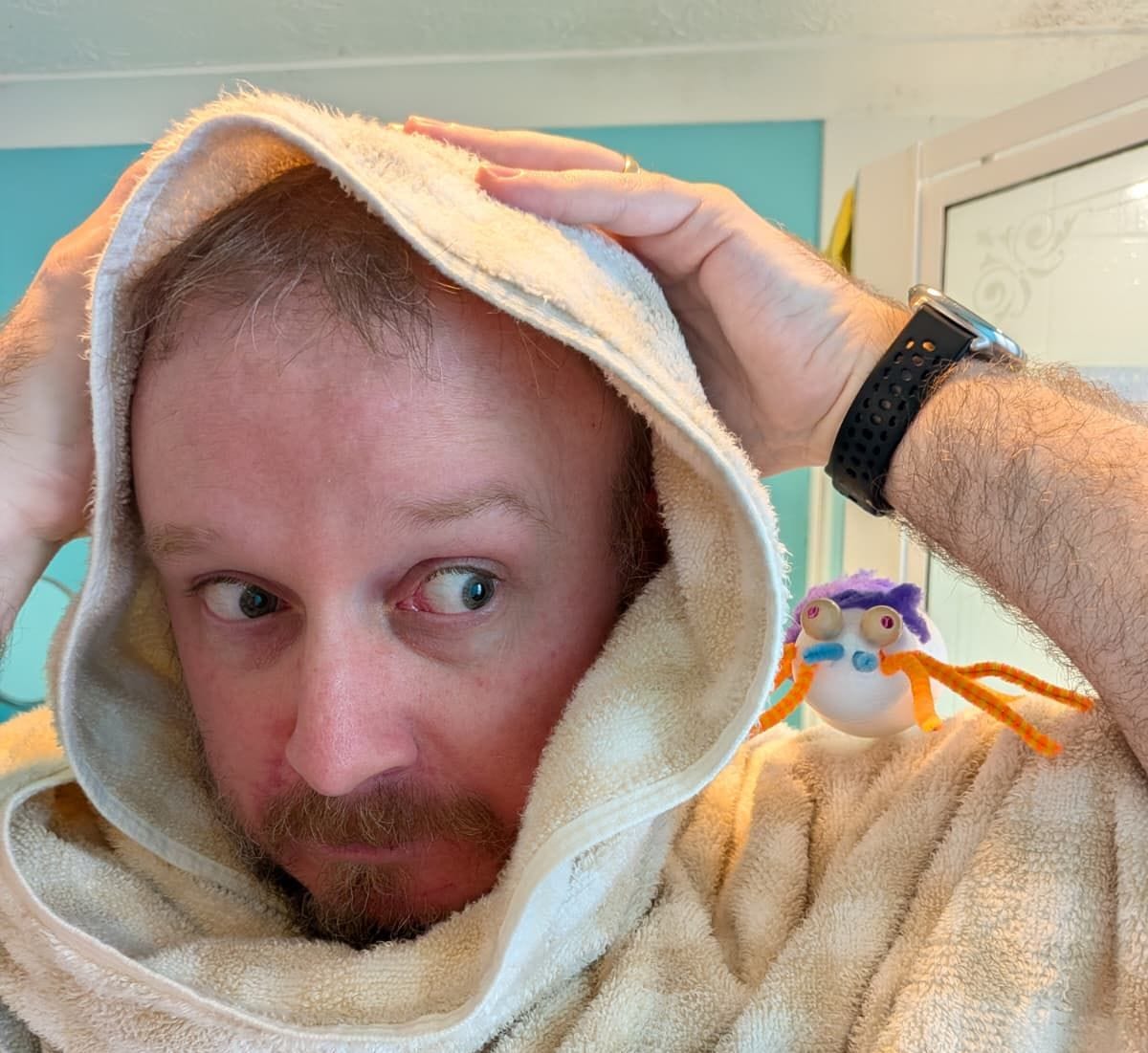 A man with facial hair dries his hair with a towel. A spider (a pretend one made from craft objects) is perched on his shoulder.