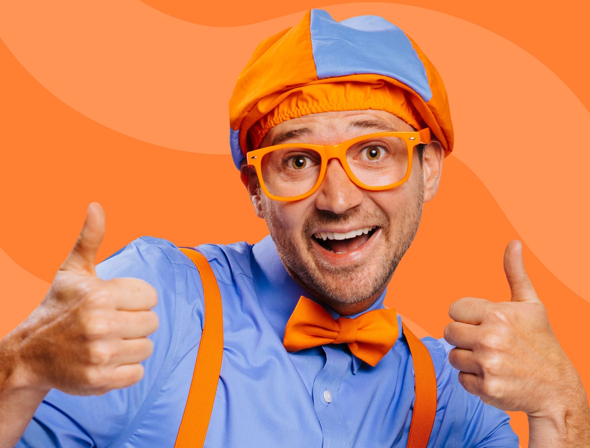 A man with short facia hair, wearing orange glasses, a blue shirt, orange bowtie, orange braces, and an orange and blue shower cap gives the camera two thumbs up.
