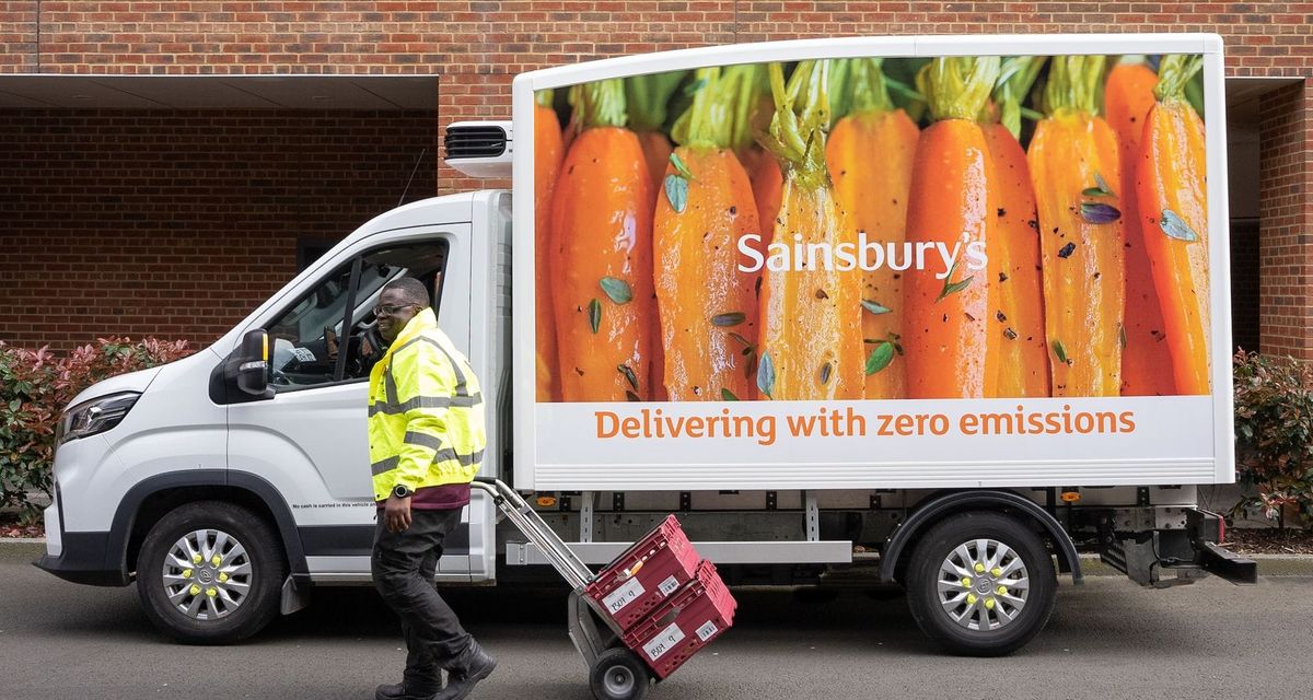 A Sainsbury's delivery truck with the driver walking alongside it pulling two crates of groceries.