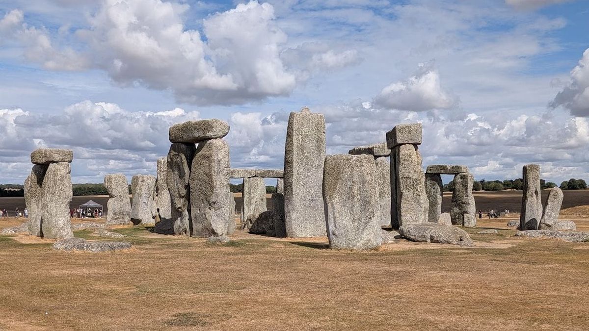 A photo of Stonehenge in the sun. The sky is blue with clouds, the ground is dry and mostly brown.