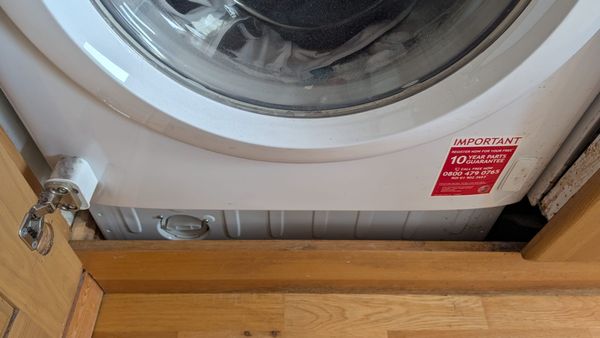 The lower half of a Hoover washing machine. The cap for the filter is visible in the lower left corner. A wooden beam covers the front of the machine on the floor.