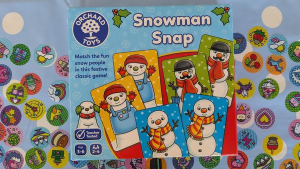 A photo of the box for the game Snowman Snap, which features a selection of happy snow people on cards. The background is a tablecloth covered in stickers.