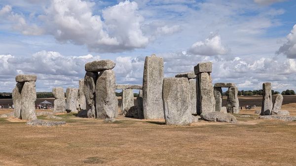 A photo of Stonehenge in the sun. The sky is blue with clouds, the ground is dry and mostly brown.
