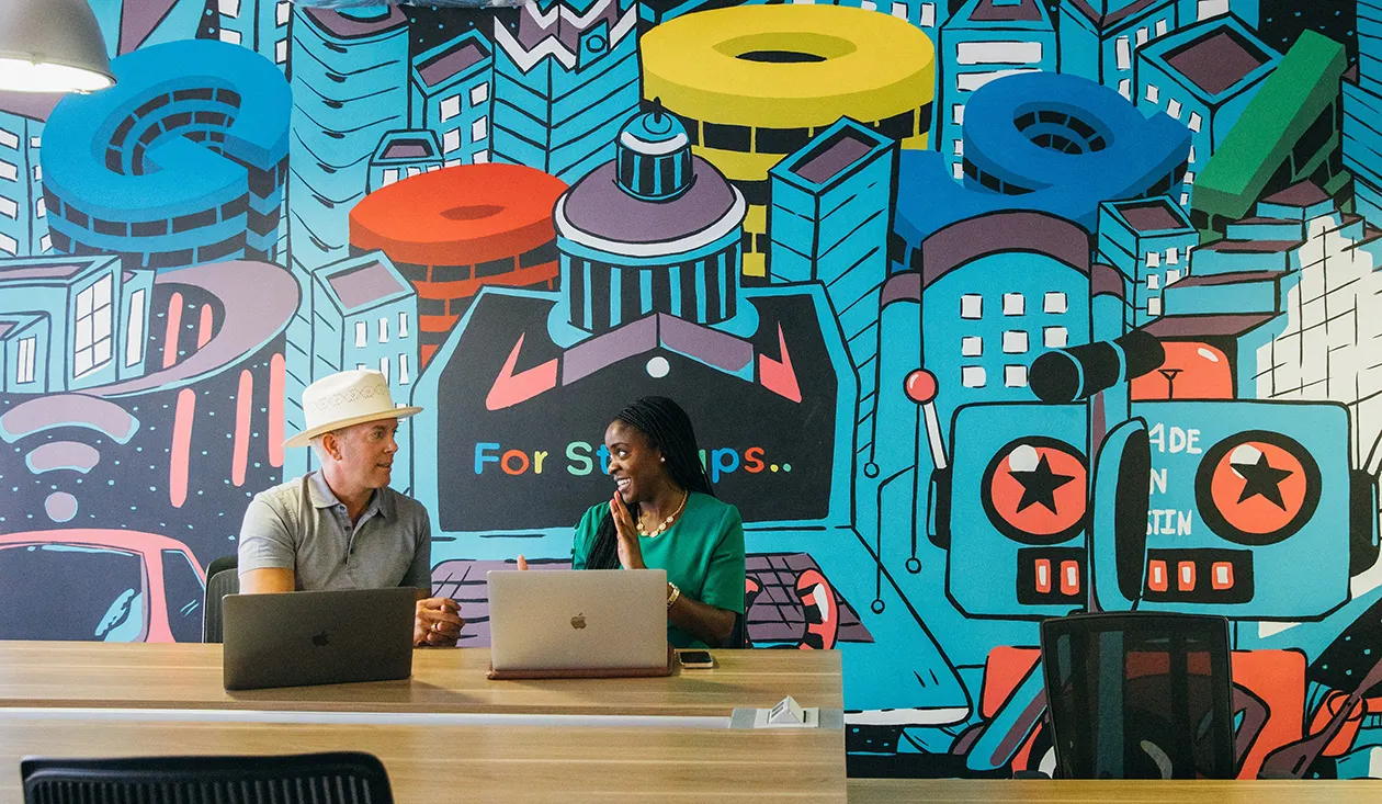 Two people sit at a desk in front of a brightly coloured Google Startups mural of buildings and a robot.