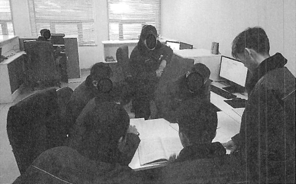 A black and white image shows a group of people with their faces blacked out around a desk. They are allegedly North Korean citizens posing as US IT workers indicted by the DoJ