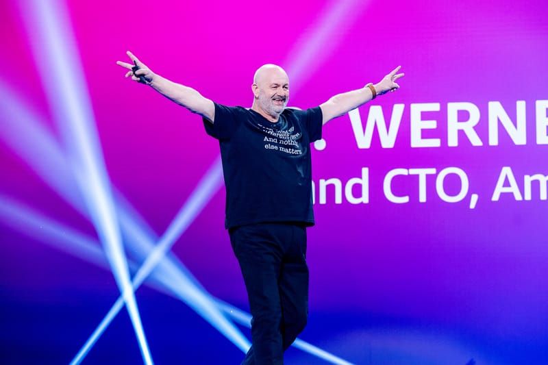 Amazon CTO Werner Vogels waves to the re:Invent 2025 crowd making two "V" gestures on stage.