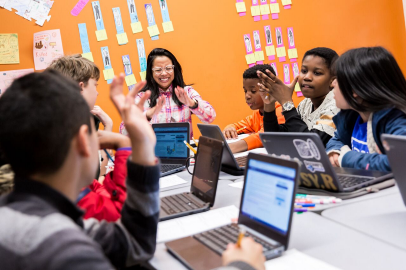 Teacher and students laugh around table with laptops open to CommonLit lesson. 