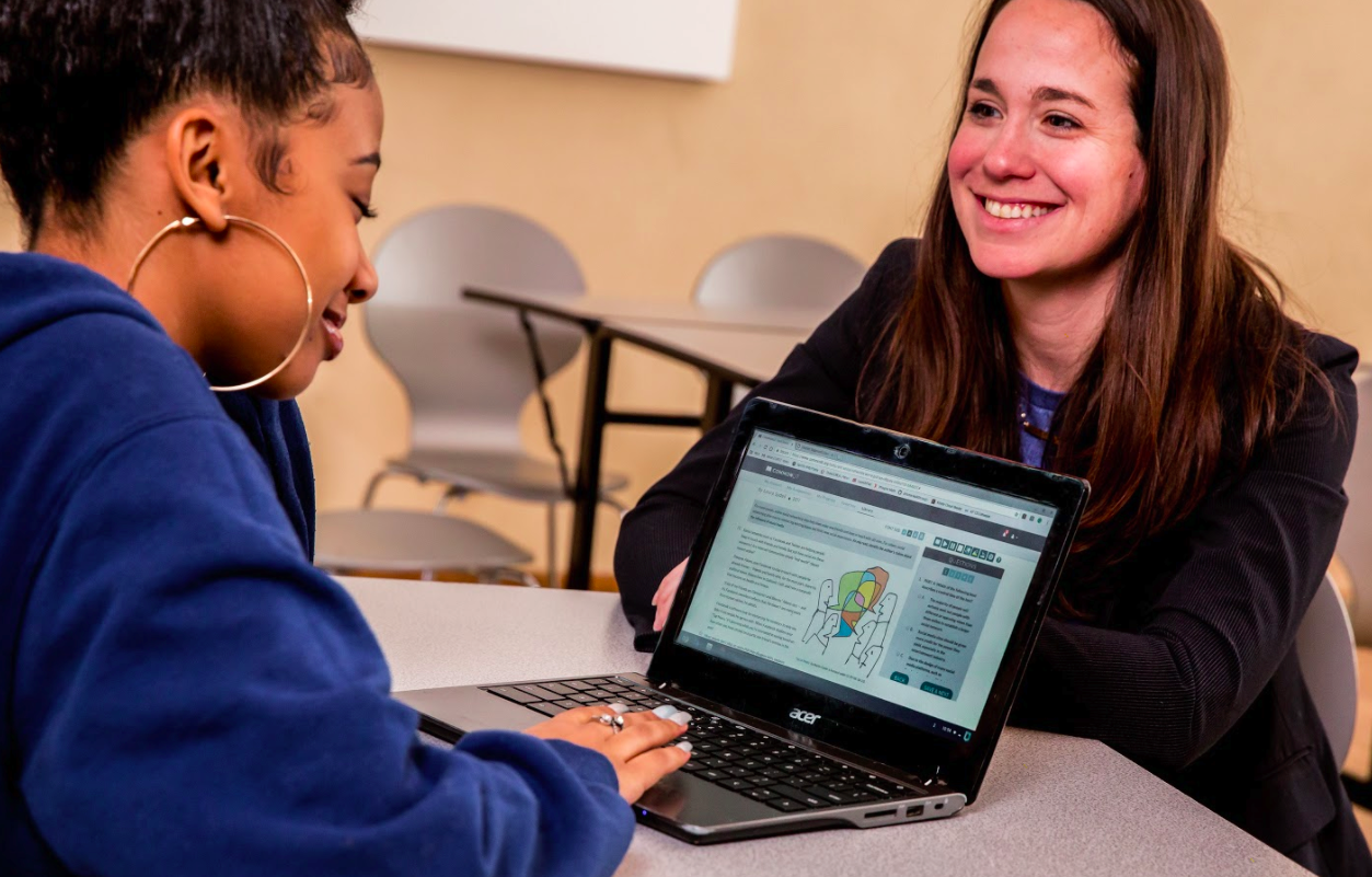 A teacher and student facing each other at a table, smiling. The student is working on a CommonLit lesson.