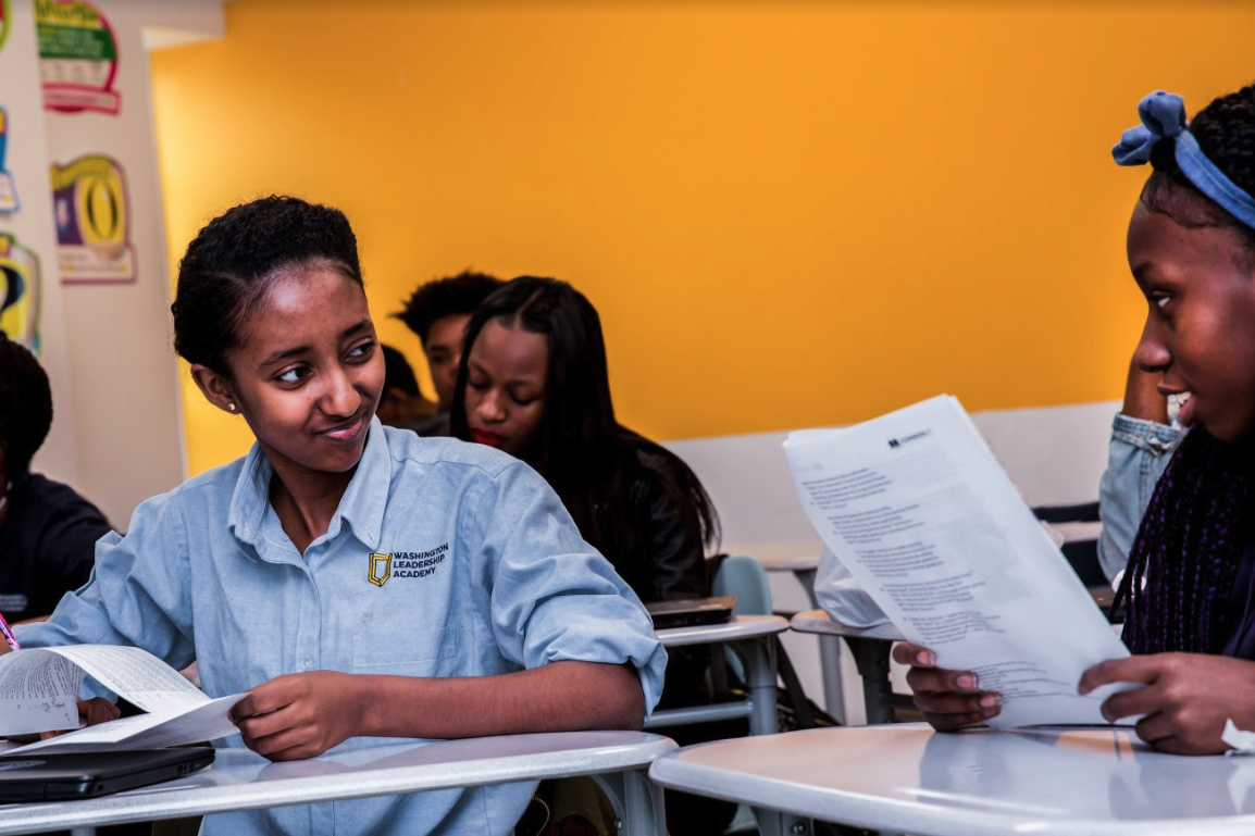 Two students sitting at desks, holding printed CommonLit lessons, and looking at each other.