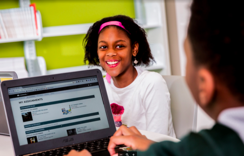 Two students sitting at a table with their laptops, facing each other.