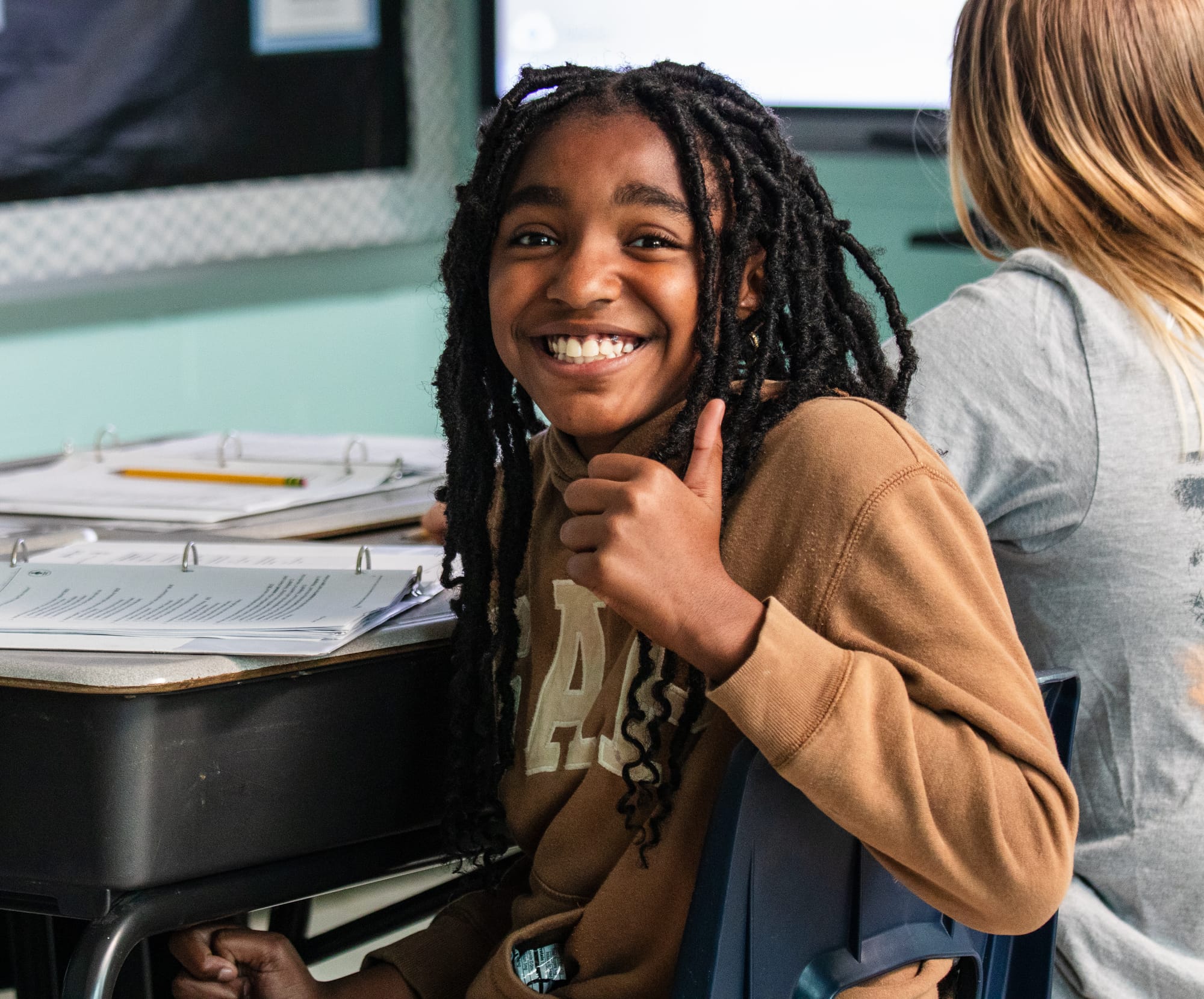 Student smiling and holding a thumbs up while completing CommonLit ELA curriculum lessons at a desk.
