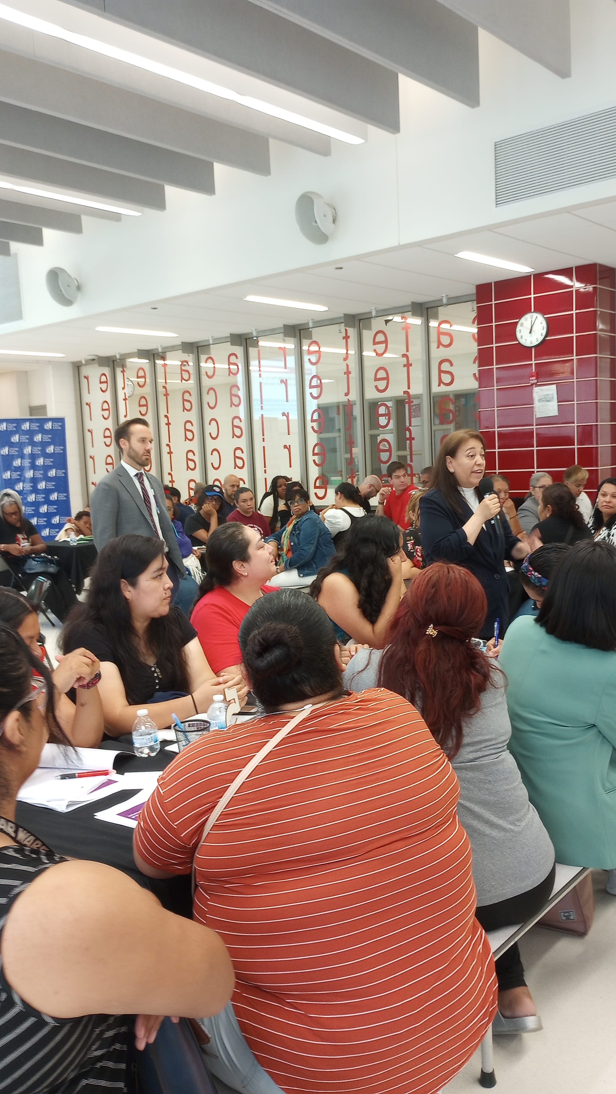A woman holds a cordless mic and speaks to a crowd seated at cafeteria tables. CPS Budget Director Mike Sitkowski stands behind the woman with the mic.