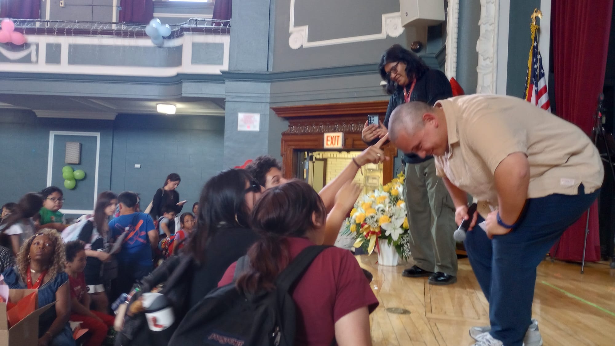 In a school auditorium, three young people reach their hands toward a man bowing at the waist. One young person's index finger touched the man's bald head.